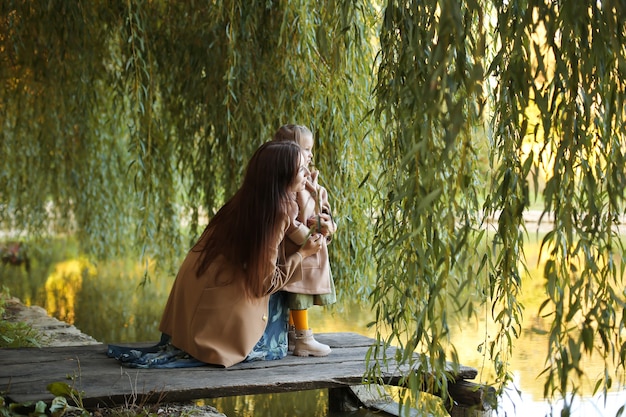 Mãe jovem de casaco abraça sua filha sentada na ponte do lago e dos ramos