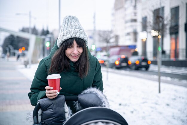 Mãe jovem e elegante com uma xícara de café em uma caminhada com um carrinho de bebê no inverno