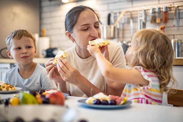 Mãe jovem feliz abraçando crianças pequenas sentindo amor e ternura sentada à mesa com sobremesa de verão caseira fresca waffles e frutas pequeno-almoço de fim de semana em família na cozinha de casa com emoção positiva
