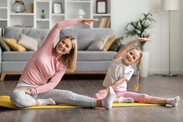 Mãe jovem sorridente e filha pequena se exercitando em um tapete de fitness em casa