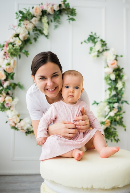 Mãe morena abraça filha bebê em fundo branco com flores