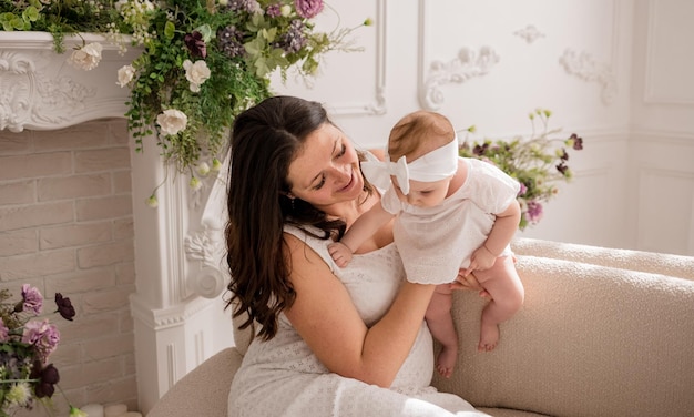 Mãe morena caucasiana está sentada no sofá e brincando com sua filha bebê em uma sala com flores