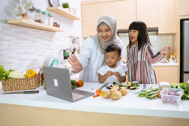 Mãe muçulmana assistindo a um vídeo de culinária no laptop e fazendo o jantar com seus dois filhos na cozinha juntos