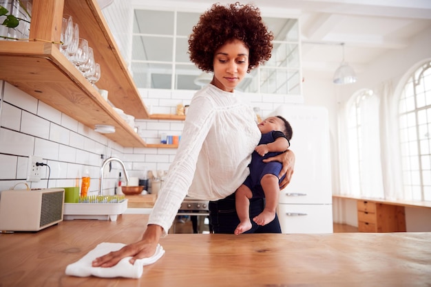 Mãe multitarefa segura filho adormecido e limpa na cozinha