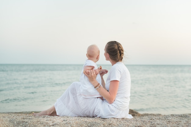 Mãe nova e criança que sentam-se na costa arenosa da parede do mar. Maternidade, harmonia e amor