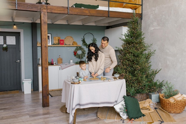 Mãe, pai e filho pequeno cortam formas de massa para biscoitos de gengibre ou uma casa de pão de gengibre na cozinha de estilo escandinavo se preparando para o Natal