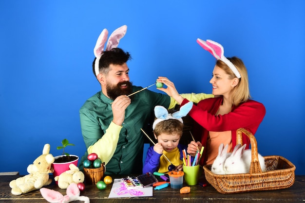 Mãe, pai e filho pintando ovos de Páscoa, criança segurando uma cesta com ovos pintados