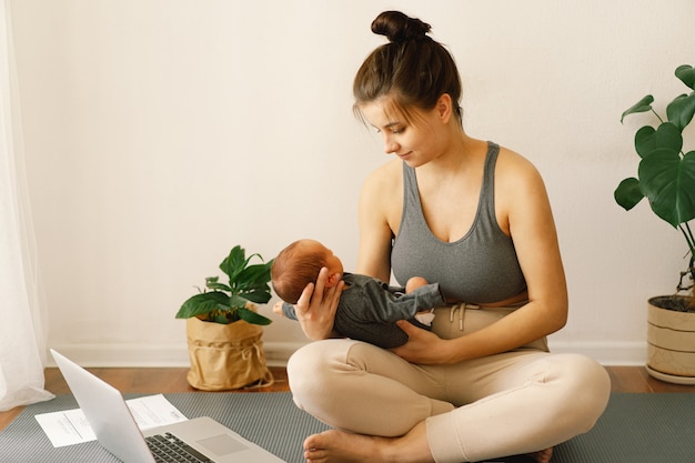 Mãe segura seu filho recém-nascido e trabalha no computador em casa