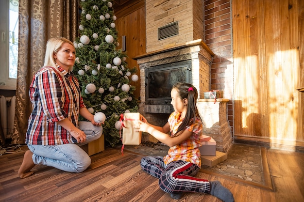 Mãe segura uma filha na árvore de Natal em uma noite festiva em uma casa de madeira rústica, ano novo, inverno