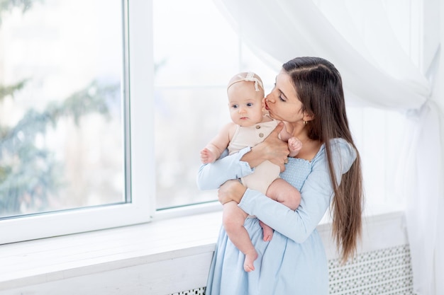 Mãe segura uma menina recém-nascida em um terno de algodão na janela da casa, abraçando-o e beijando-o, feliz maternidade ou família