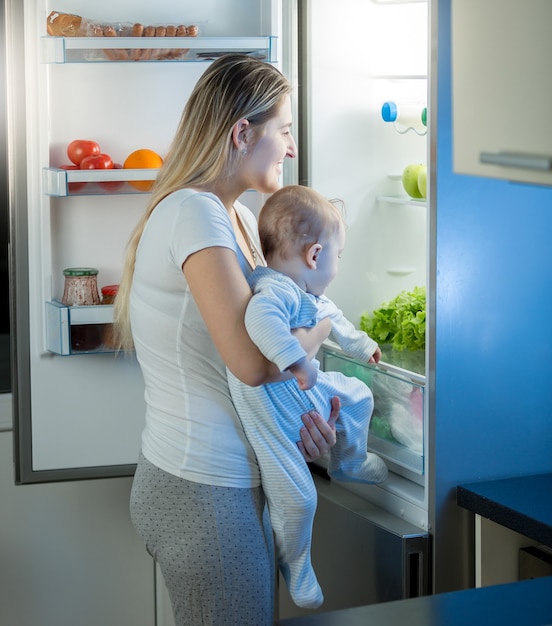 Mãe segurando filho bebê e olhando dentro da geladeira à noite