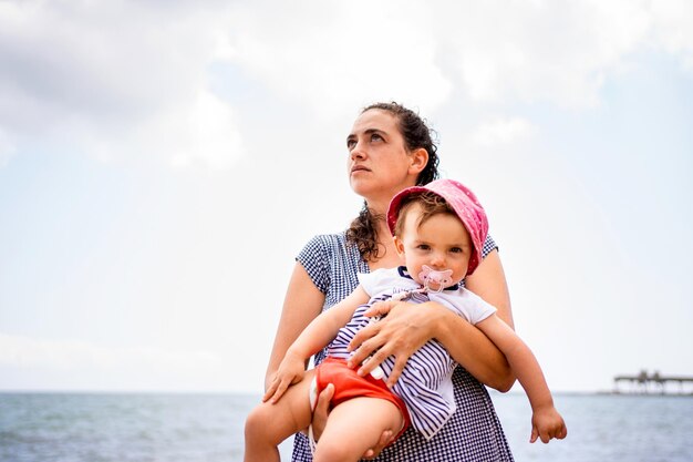 Mãe segurando um bebê nos braços enquanto olha para o céu ao lado do oceano