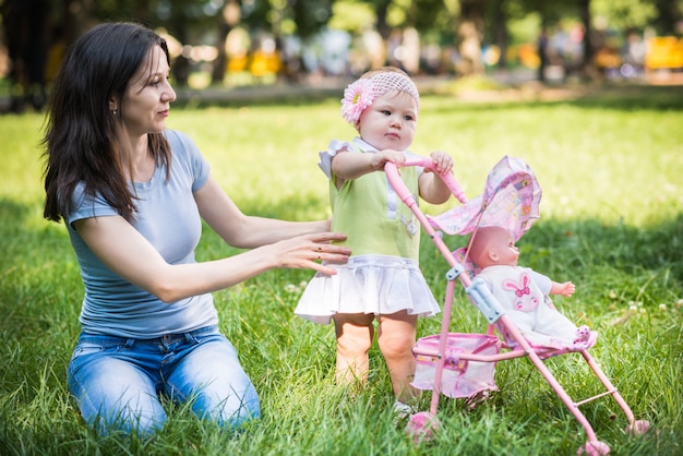 Mãe sentada na grama e ajudando a filha a andar e rolar carrinho de brinquedo