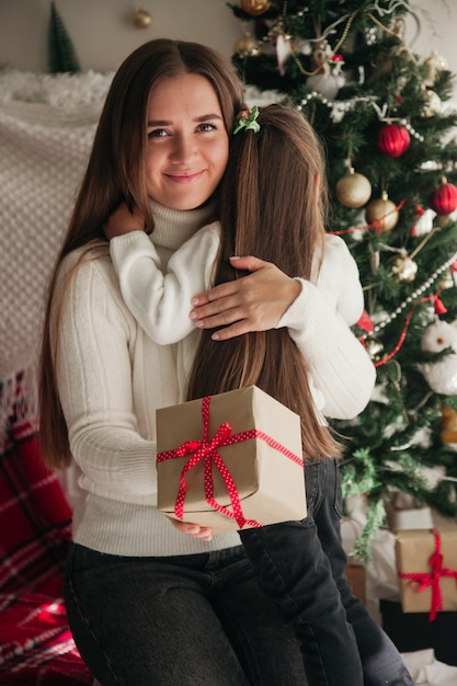 Mãe sorridente abraçando a filha com presentes nas mãos e decorações de natal ao fundo conceito de presentes de família de natal