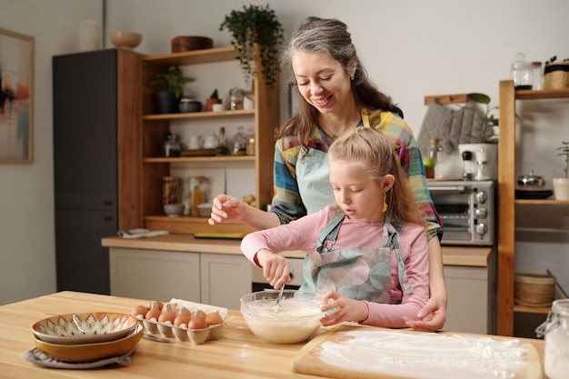 Mãe sorridente ajudando a filha na cozinha enquanto faz bolo