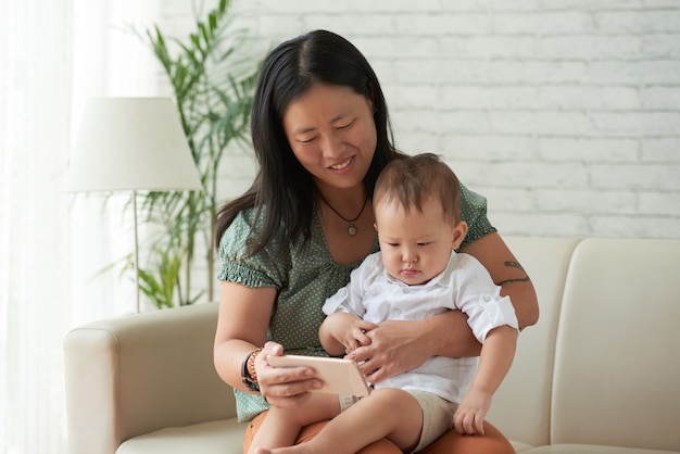 Foto mãe sorridente mostrando desenho animado ou vídeo educacional no smartphone para o filho pequeno sentado no sofá em casa