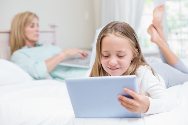 Mãe usando o laptop enquanto a filha usa tablet pc na cama