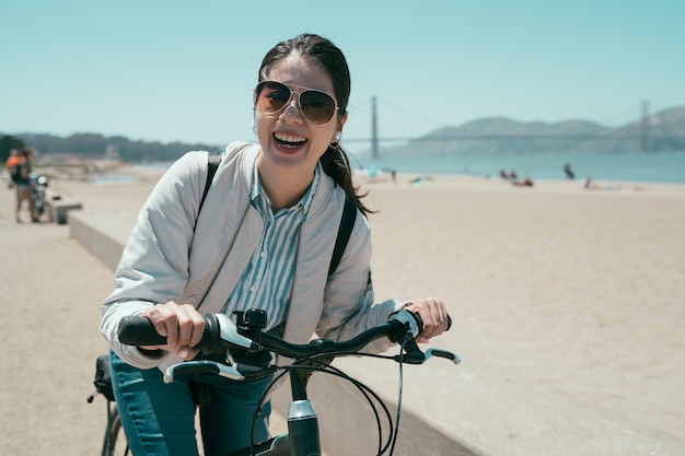 Mädchen Backpacker mieten Fahrradreise Tour in San Francisco Kalifornien USA. junge asiatische frau gesichtskamera lächelt fröhlich, während sie fahrrad am strand fährt. Golden Gate Bridge mit blauem Himmel im Hintergrund.