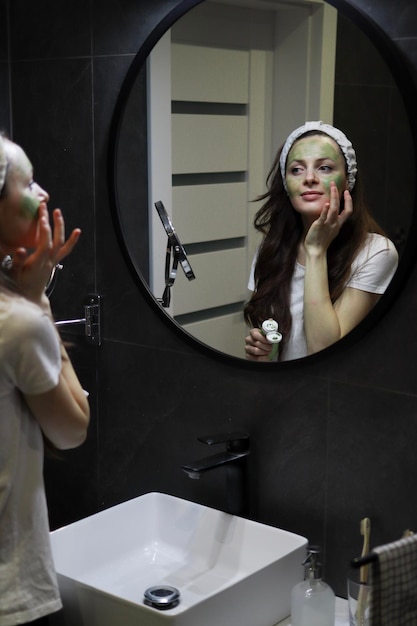 Mädchen bereitet die Haut vor einem wichtigen Ereignis vor Natürliche Hautpflege Beauty Home Spa Frau mit Stirnband, die in einem Loft-Badezimmer vor einem Spiegel eine grüne Tonmaske auf ihr Gesicht aufträgt