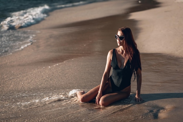 Mädchen Brunette in einem schwarzen Badeanzug und einer schwarzen Sonnenbrille am Strand mit weißem Sand in der Nähe des Ozeans im Urlaub. Ein schönes Modell mit einem sexy Körper sonnt sich im Sand am Strand.Thailand.Phuket