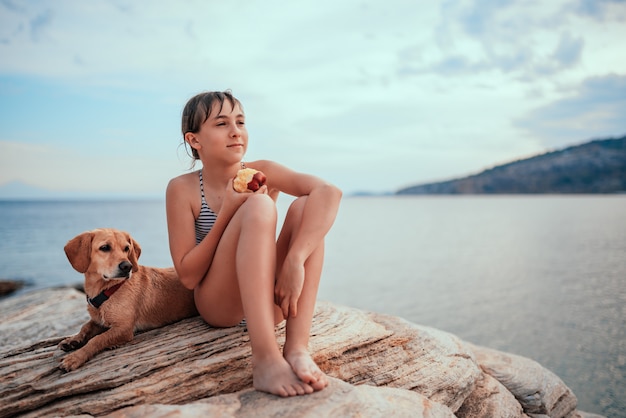 Mädchen, das am felsigen Strand mit Hund sitzt und Apfel isst