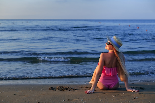 Mädchen, das am Strand im rosa Badeanzug und im Bootsfahrer sitzt.