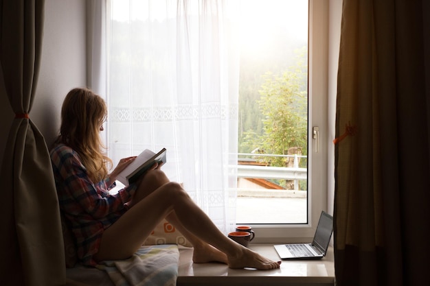 Mädchen, das ein Buch hält, das auf einer Fensterbank sitzt, mit hellem Sonnenlicht aus dem Fenster