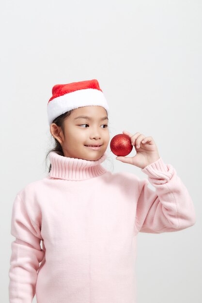 Mädchen, das eine rote Weihnachtskugel in der Strickjacke und im Sankt-Hut anhält