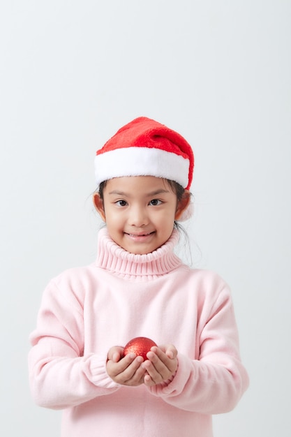 Mädchen, das eine rote Weihnachtskugel in der Strickjacke und im Sankt-Hut anhält