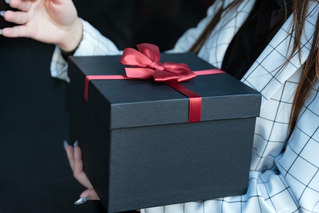 Mädchen, das eine schöne Geschenkbox in ihren Händen hält. Blackbox mit roter Schleife in weiblichen Händen auf schwarzem Hintergrund. Platz kopieren