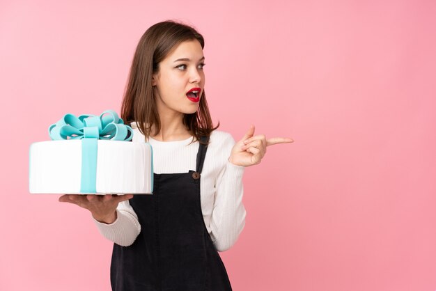 Mädchen, das einen großen Kuchen lokalisiert auf rosa Wand überrascht hält und Finger zur Seite zeigt