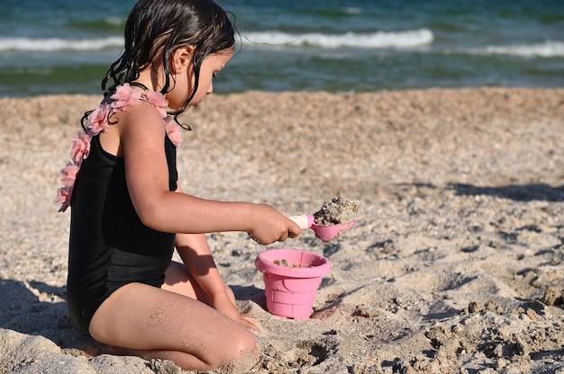 Mädchen, das einen Rechen hält und den rosa Spielzeugeimer mit Sand füllt, der sandige Formen und Burgen baut