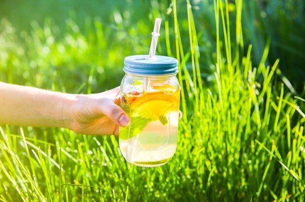 Mädchen, das frische Limonade in Gläsern mit Strohhalmen hält. Hipster-Sommergetränke. Umweltfreundlich in der Natur. Zitronen, Orangen und Beeren mit Minze im Glas. Grünes hohes Gras draußen.