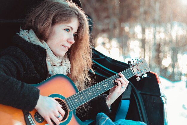 Mädchen, das Gitarre spielt, das auf dem Kofferraum des Autos im Winterwald sitzt - Reisen mit dem Auto