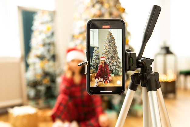 Foto mädchen, das in der nähe des weihnachtsbaums sitzt und ihren weihnachtsvideoblog mit smartphone und stativ dreht. sie trägt einen roten pyjama und eine weihnachtsmannmütze.