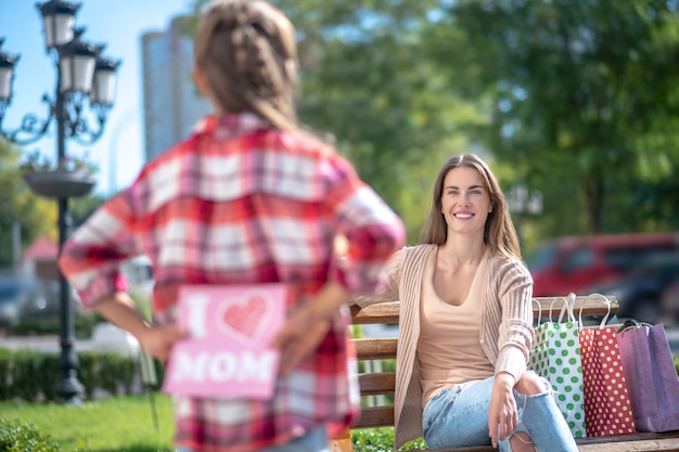 Foto mädchen, das karte für ihre lächelnde mutter hält, die auf parkbank sitzt