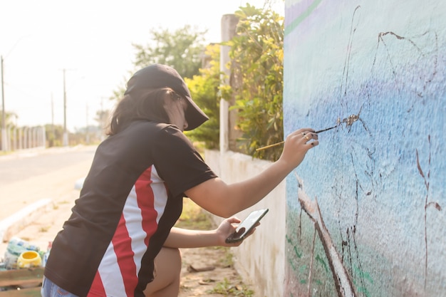 Mädchen, das sich darauf konzentriert, eine Straßenmauer mit ihrem Handy als Referenz zu malen.