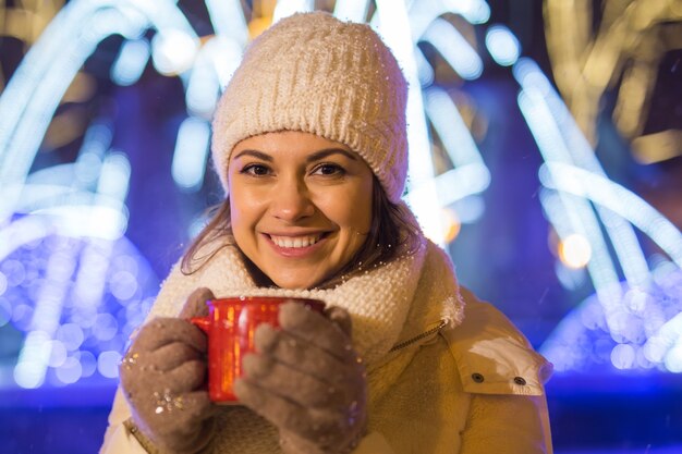 Mädchen, das über Winterweihnachtsstadthintergrund Schneeschneeverwehungen steht, steht warme Jacke Hut hält