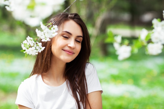 Mädchen, das unter blühendem Apfelbaum im Naturpark steht. Modekonzept. Elegante junge Dame in der Jeanskleidung, die blühenden Garten am sonnigen Frühlingstag genießt. Natürliches Schönheits-Make-up. Horizontal