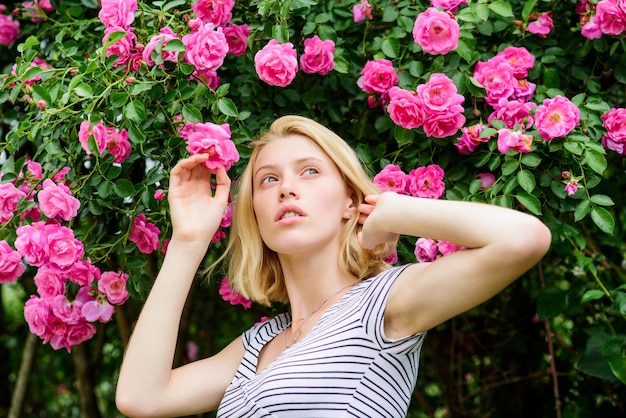 Mädchen entzückender blonder schnüffelnder Duft der rosa Blüte. Frühling und Sommer. Parfüm und Kosmetik. Frau vor blühendem Rosenbusch. Blüte von Wildrosen. Geheimes Gartenkonzept. Duft nach Rosen.