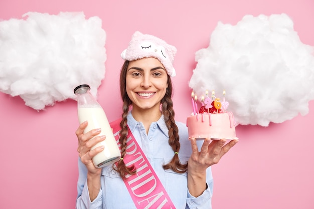 Mädchen feiert 26. Geburtstag hält festlichen Kuchen mit brennenden Kerzen und eine Flasche Milch, die ein leckeres Frühstück haben wird, trägt Schlafmaske und Hemd isoliert auf rosa Wand