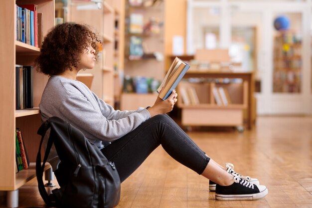 Mädchen gemischter Abstammung in der Freizeitkleidung, die auf dem Boden durch Bücherregale in der Universitätsbibliothek sitzt und Buch liest