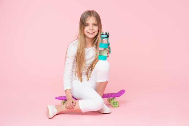 Mädchen glückliches Gesicht hält mit Wasserflasche, während Penny Board rosa Hintergrund sitzen Kind Mädchen Pflege Körper Hydratation Aktive Freizeit und Wasserhaushalt Aktives und gesundes Kind trinkt Wasser Bleiben hydratisiert