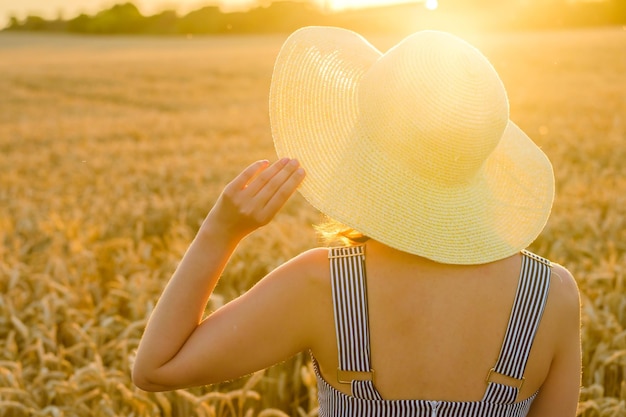 Mädchen hält sich an Strohhut fest, der bei Sonnenuntergang im Weizenfeld steht