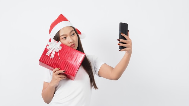 Mädchen halten Geschenkbox machen Selfie oder Video online mit Weihnachten Weihnachtsstütze Dekoration. Asiatische thailändische jugendlich Frau, die Online-Selfie nimmt, um festliche Jahreszeit mit ihrem Freund durch rote Geschenkbox zu feiern. Studioaufnahme.