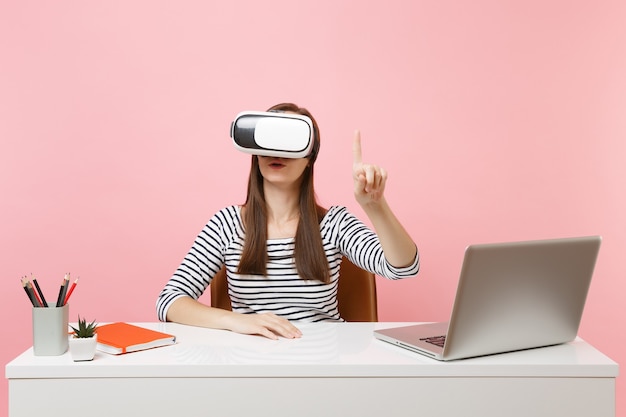 Mädchen im Headset der virtuellen Realität auf dem Kopf berühren so etwas wie Knopfdruck oder zeigen auf schwebende virtuelle Bildschirmarbeit am Schreibtisch mit Laptop einzeln auf rosafarbenem Hintergrund. Erfolg Geschäftskarriere.