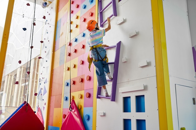 Mädchen im Helm hängt am Seil an der Kletterwand, Unterhaltungszentrum, junger Kletterer. Kinder haben Spaß, Kinder verbringen das Wochenende auf dem Spielplatz, glückliche Kindheit