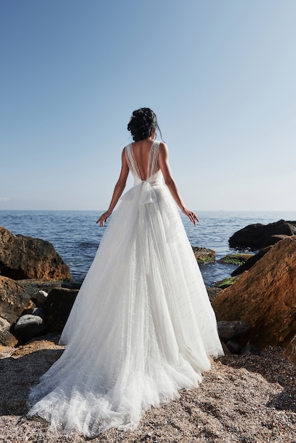 Mädchen im Hochzeitsluxuskleid, das am Seeufer aufwirft. Braut auf einem Felsen.