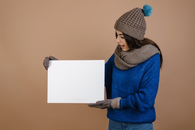 Mädchen im Hut und in Handschuhen, die weißes leeres Plakat halten. Isoliert auf braunem hintergrund.
