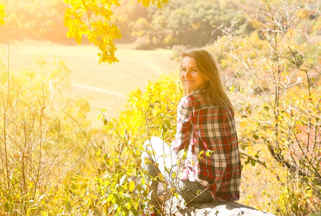 Mädchen im karierten Hemd sitzt im Herbstwald. Saisonales Konzept. Stilvolle Hipster-Kleidung im Freien. Naturphilosophie herum. Schöne Frau in der Nähe der gelben Blätter und Bäume.